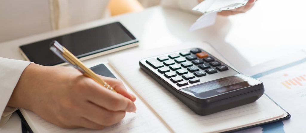 Foto de uma mulher fazendo algumas contas, usando uma calculadora e um papel.
