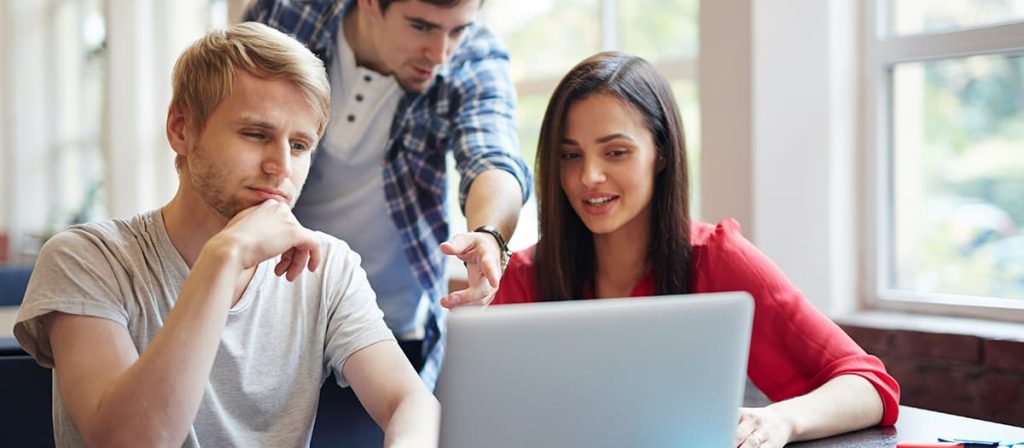 Três pessoas em uma mesa com um notebook, uma está apontando para algo no notebook.